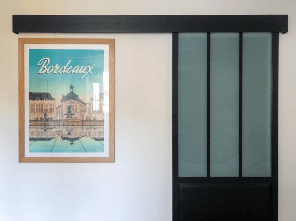 Une affiche de Bordeaux encadrée de bois, à côté de la porte vitrée noire coulissante du studio