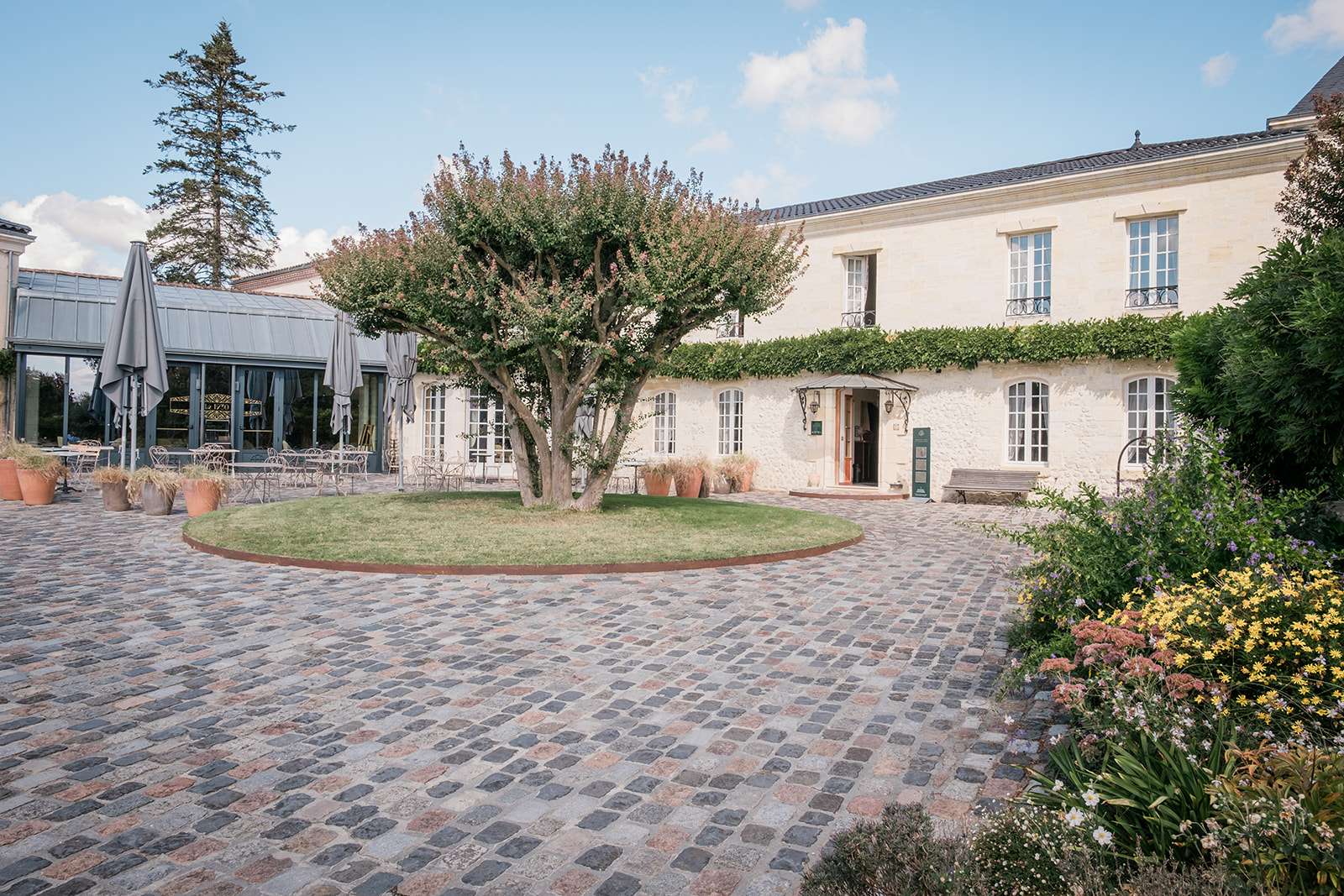 La cour pavée du Domaine de Larchey, avec son arbre au centre d'une pelouse ronde, la façade en pierre blonde et la verrière du restaurant