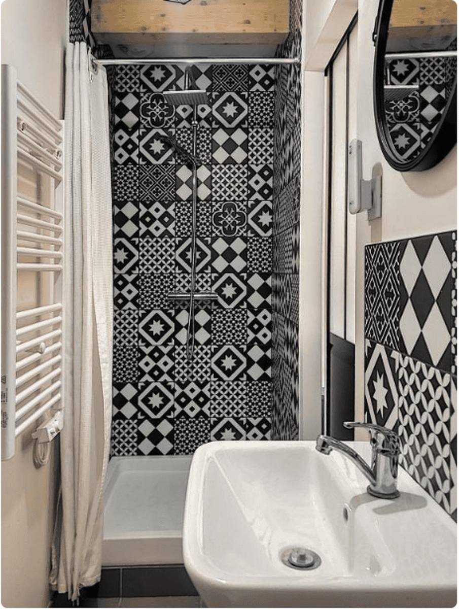 La salle de bains : douche aux carreaux de ciment noirs et blancs, lavabo blanc, sèche-serviettes et miroir rond
