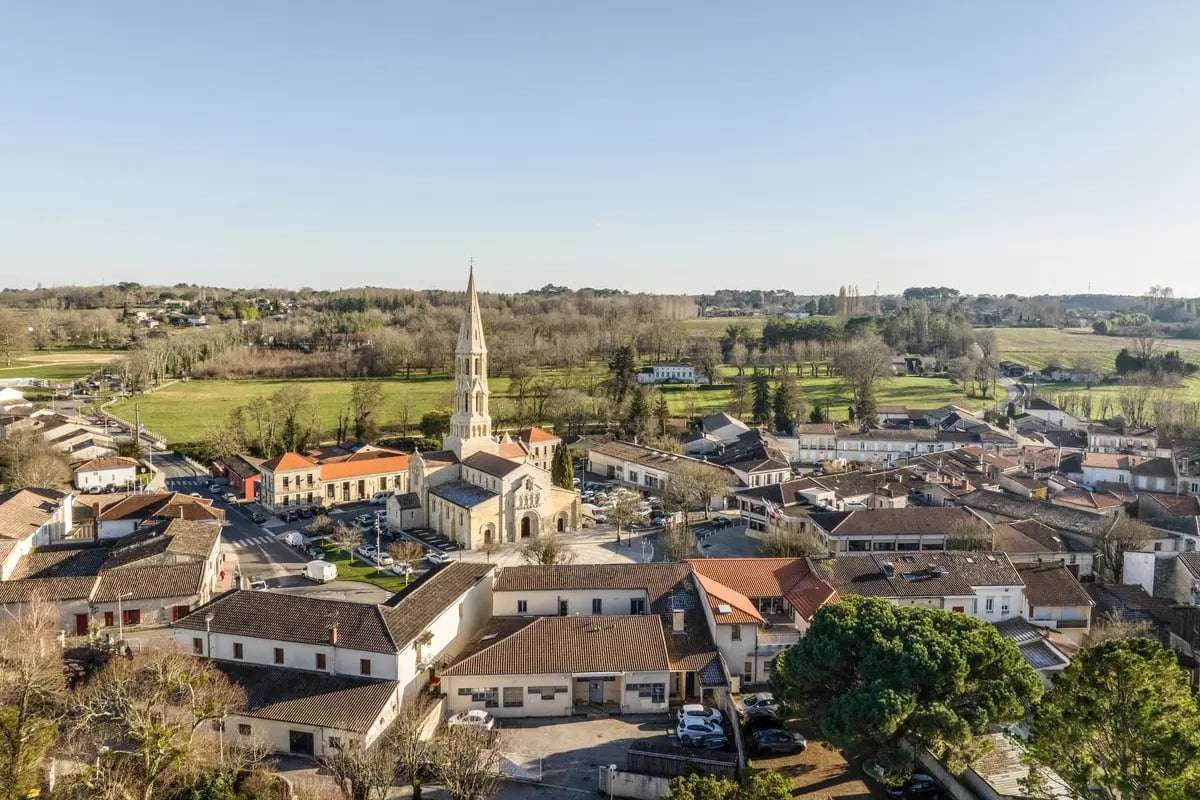 Vue aérienne du centre de La Brède un jour d'hiver ensoleillé : l'église Saint-Jean-d'Étampes et son clocher, les toits de tuile autour de la place, des prés et des bois à l'horizon.